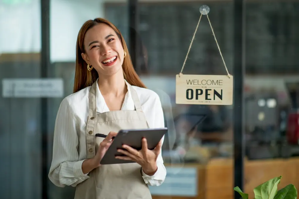 Smiling female small business owner wearing an apron and holding a digital tablet in front of her shop