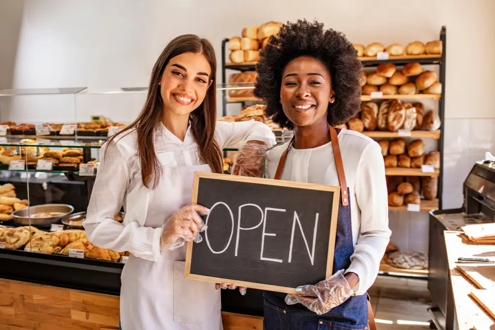 Two smiling female bakers holding an "OPEN" sign inside a bakery with shelves full of fresh bread