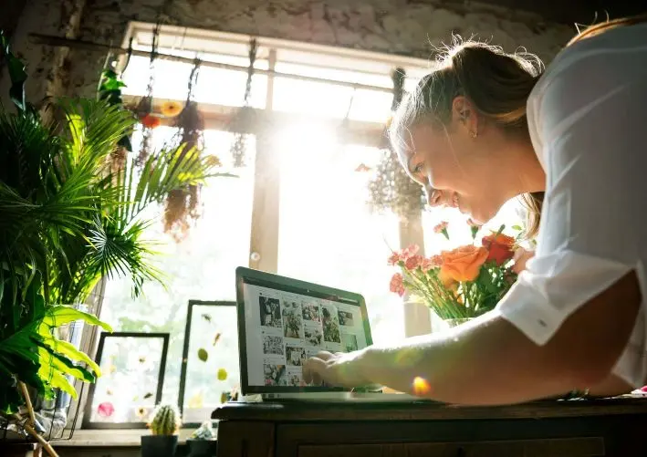 Woman browsing photos on a laptop in a sunlit room filled with plants and flowers