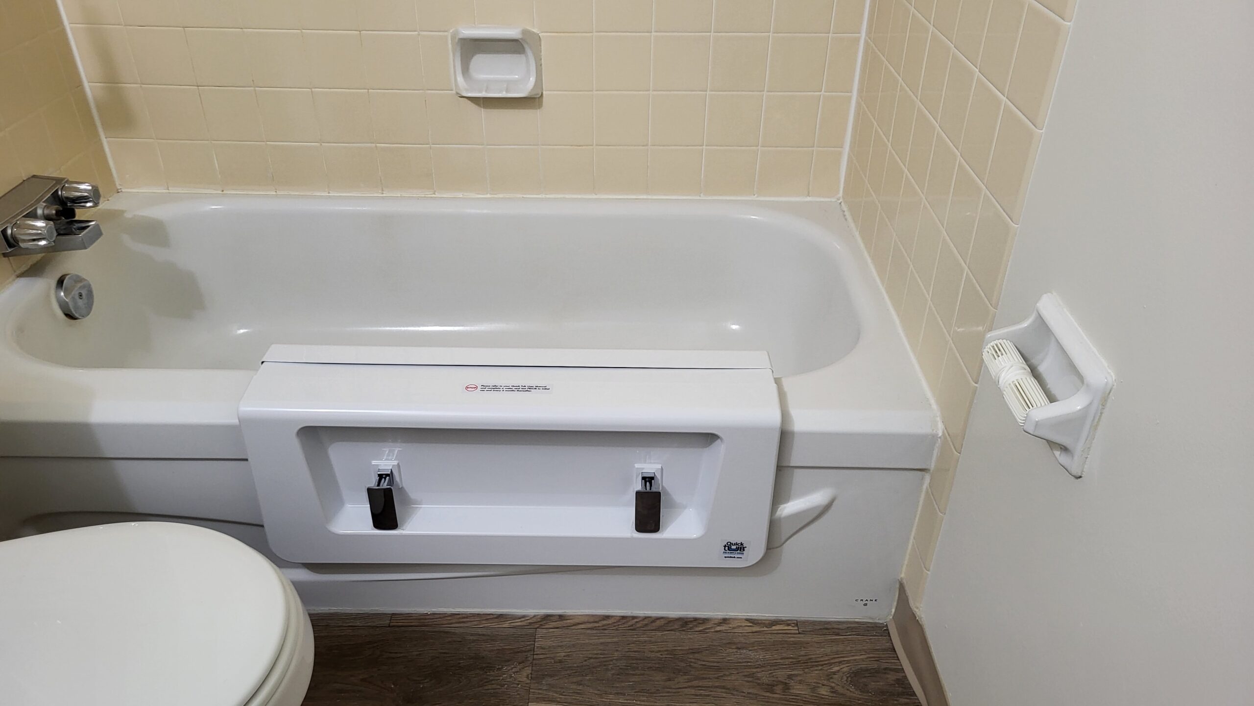 Bathtub modified with an easy-access entry panel, surrounded by beige tiles and wood-style flooring