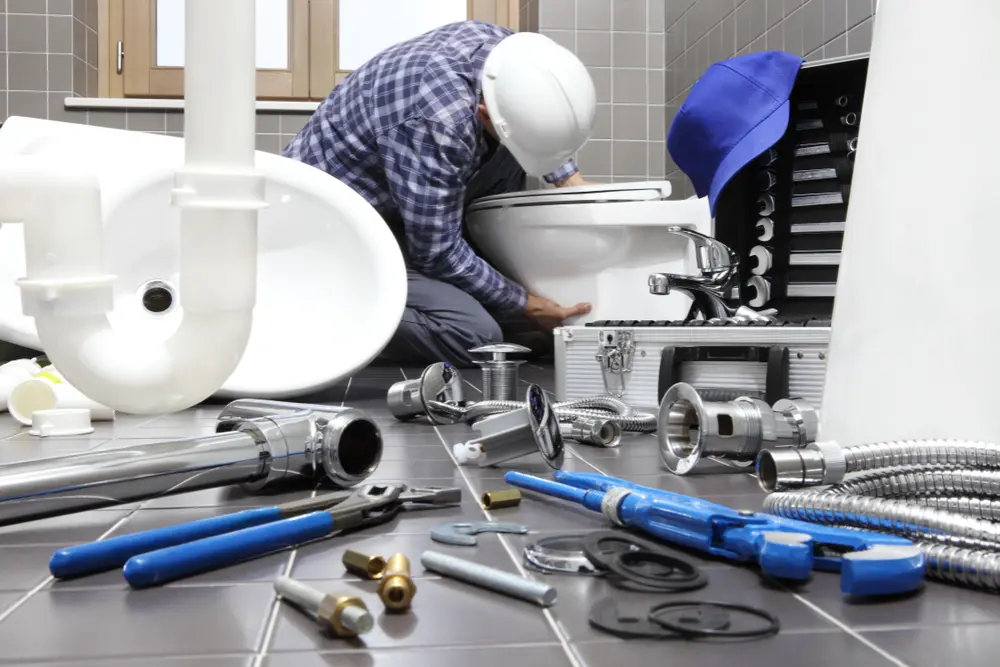 Plumber in safety gear installing a toilet amid plumbing tools on a tiled bathroom floor