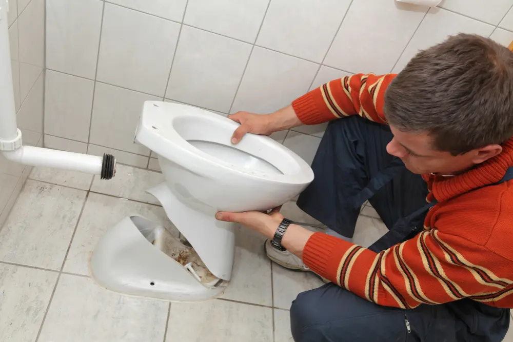 Man installing a toilet bowl onto a damaged toilet base in a tiled bathroom