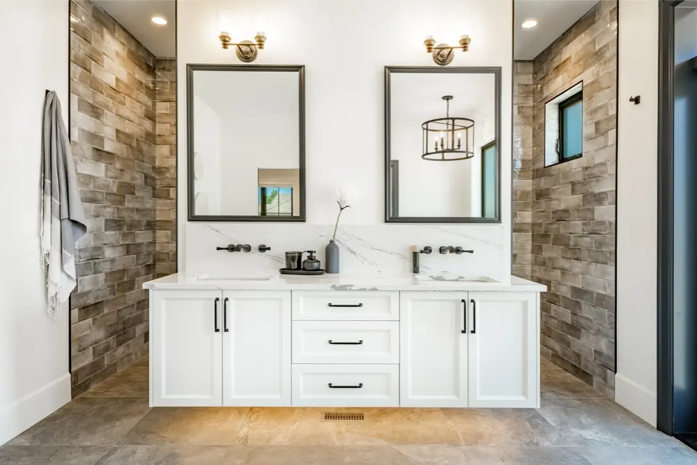 Modern double vanity bathroom with white cabinets, wall-mounted faucets, and rustic tiled walls