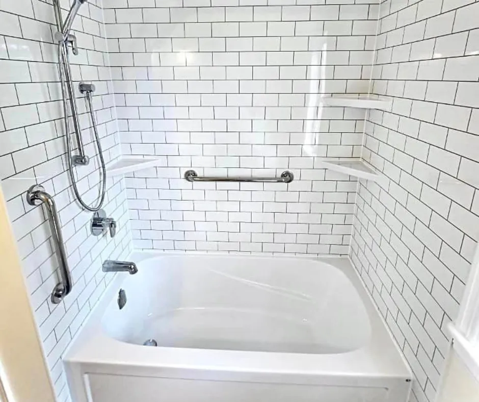 Accessible bathtub with white subway tile walls, stainless steel grab bars, and built-in corner shelves