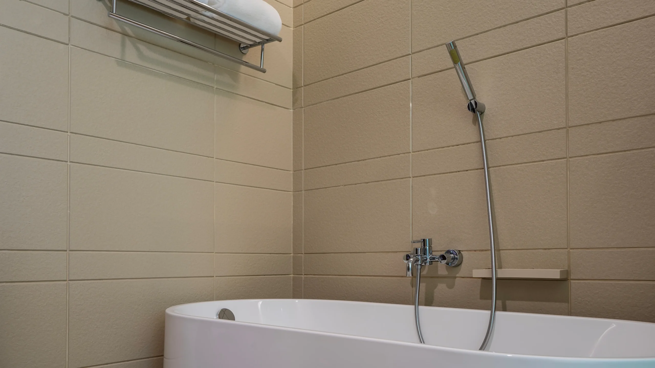 Modern tub and handheld shower with beige tiles and towel shelf