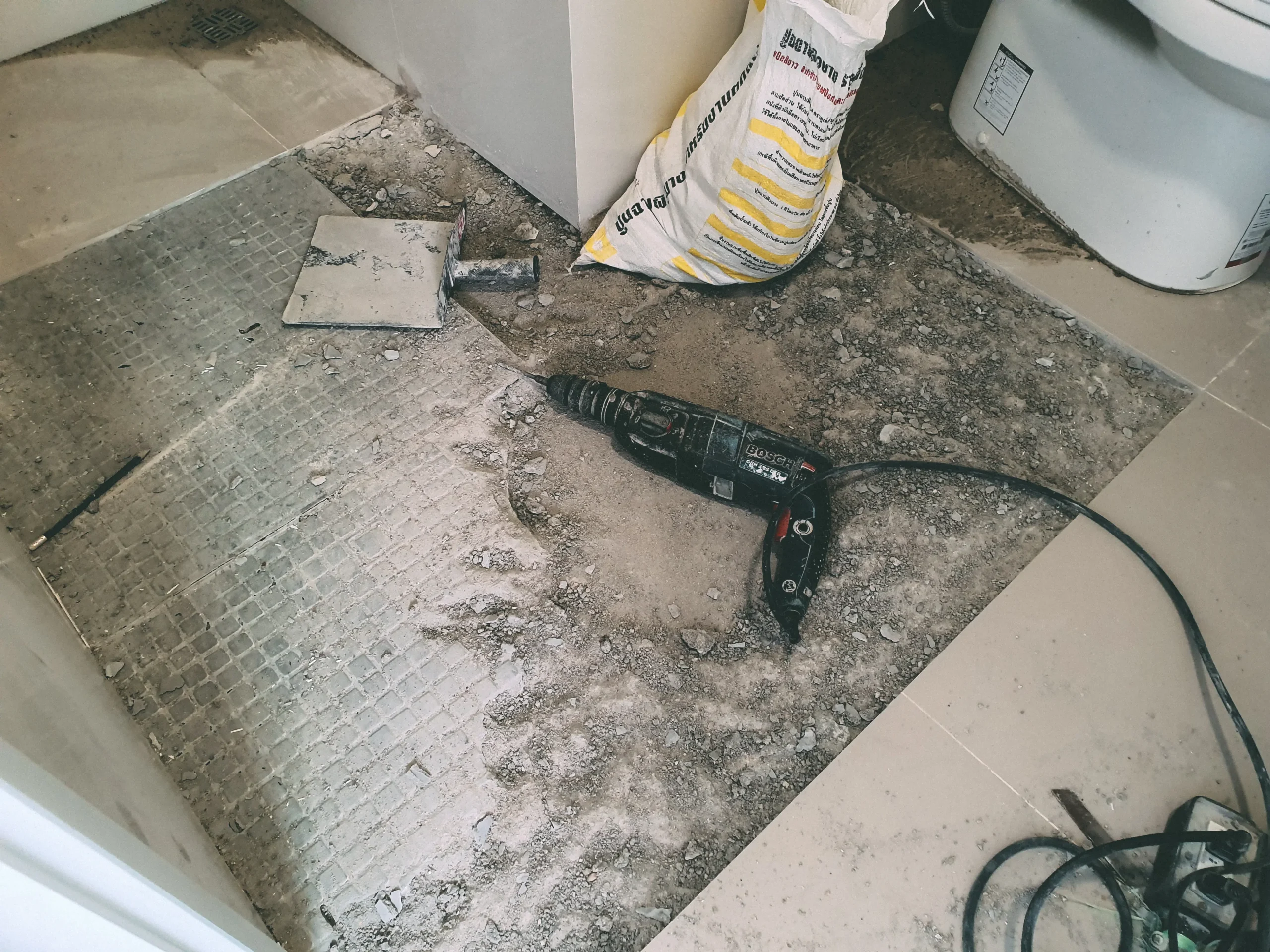 Bathroom floor during tile removal with dust, debris, and a Bosch power drill on the ground.