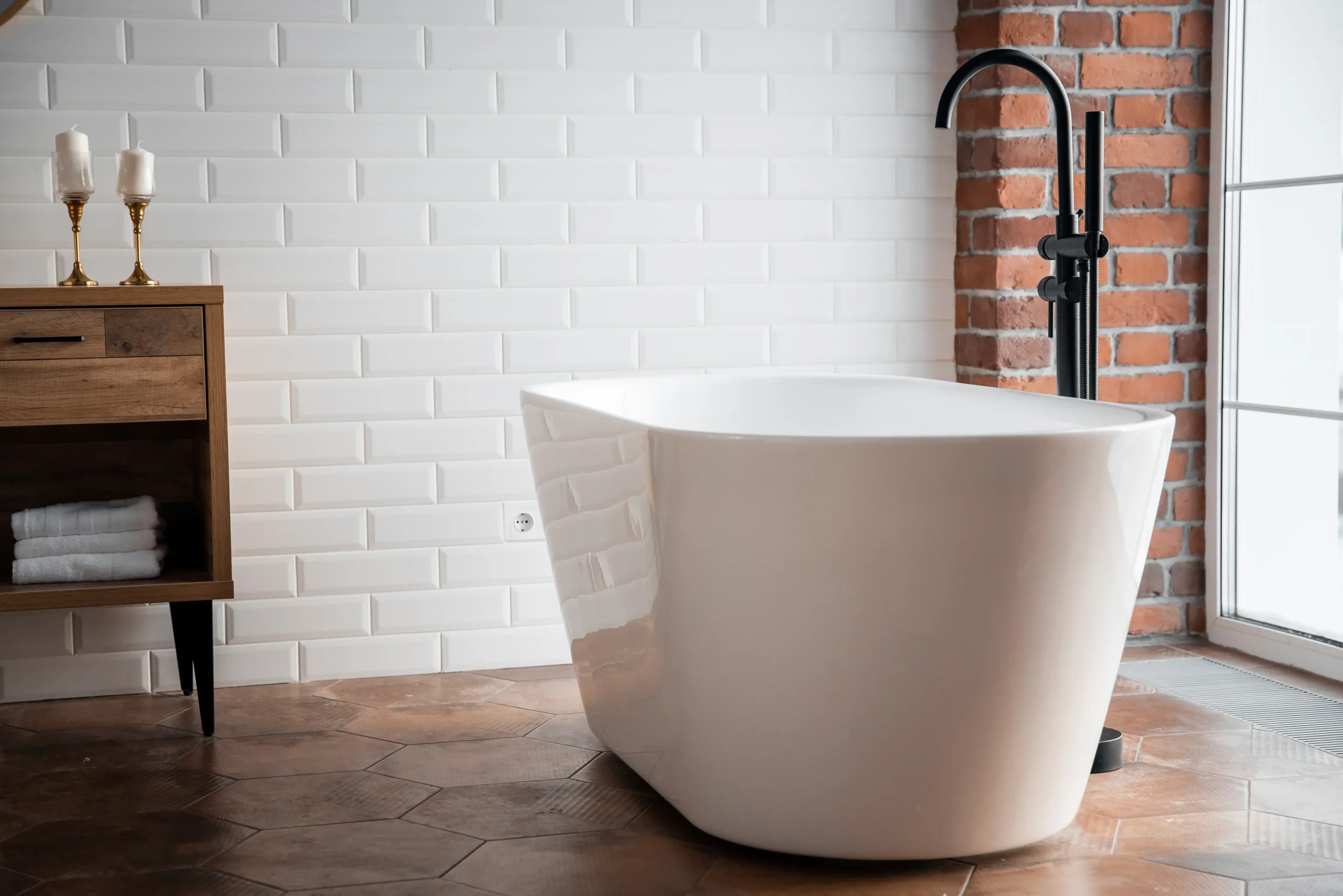 Freestanding white bathtub with black faucet in modern bathroom with white subway tile and brick walls