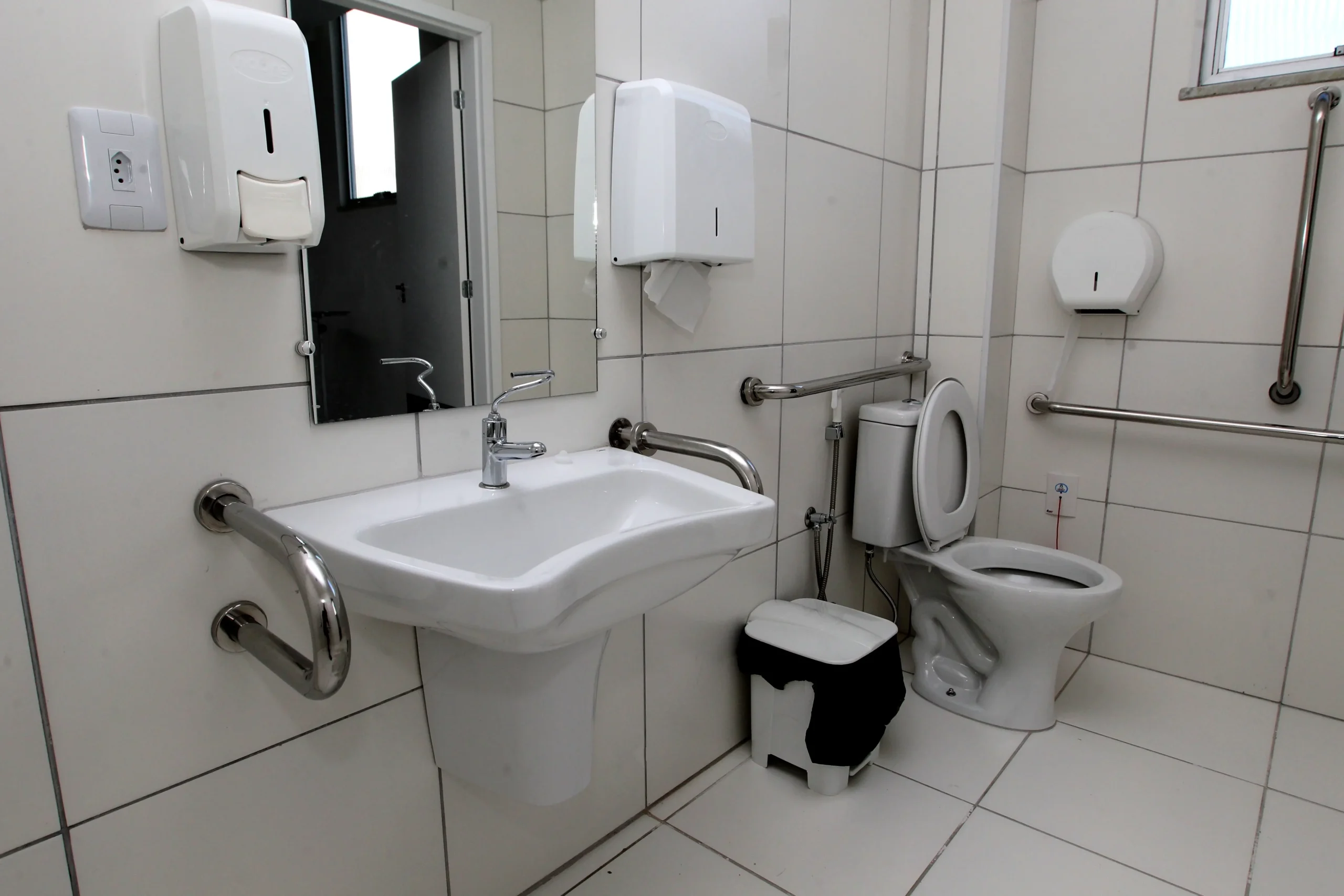 Accessible restroom with grab bars, wall-mounted sink, and toilet on white tiled walls