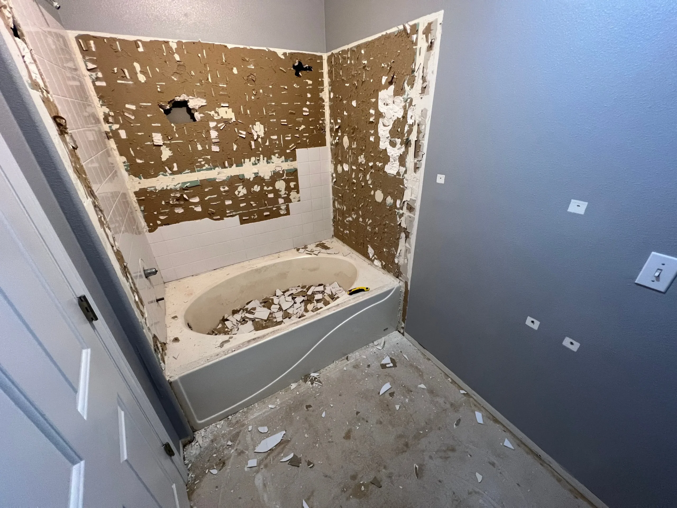 Bathroom under renovation with tile debris and exposed drywall around bathtub