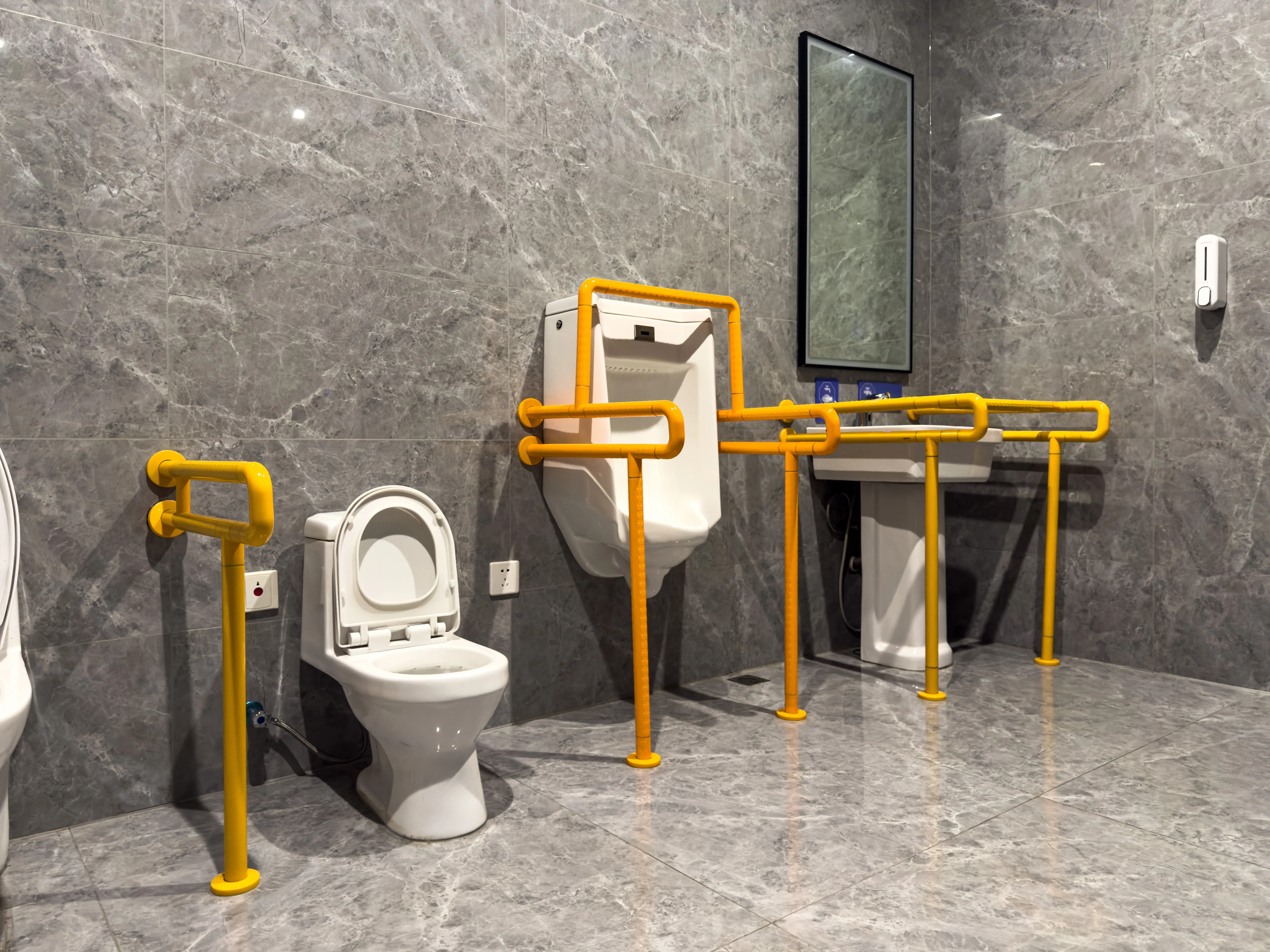 Accessible public restroom with toilet, urinal, sink, and yellow safety grab bars